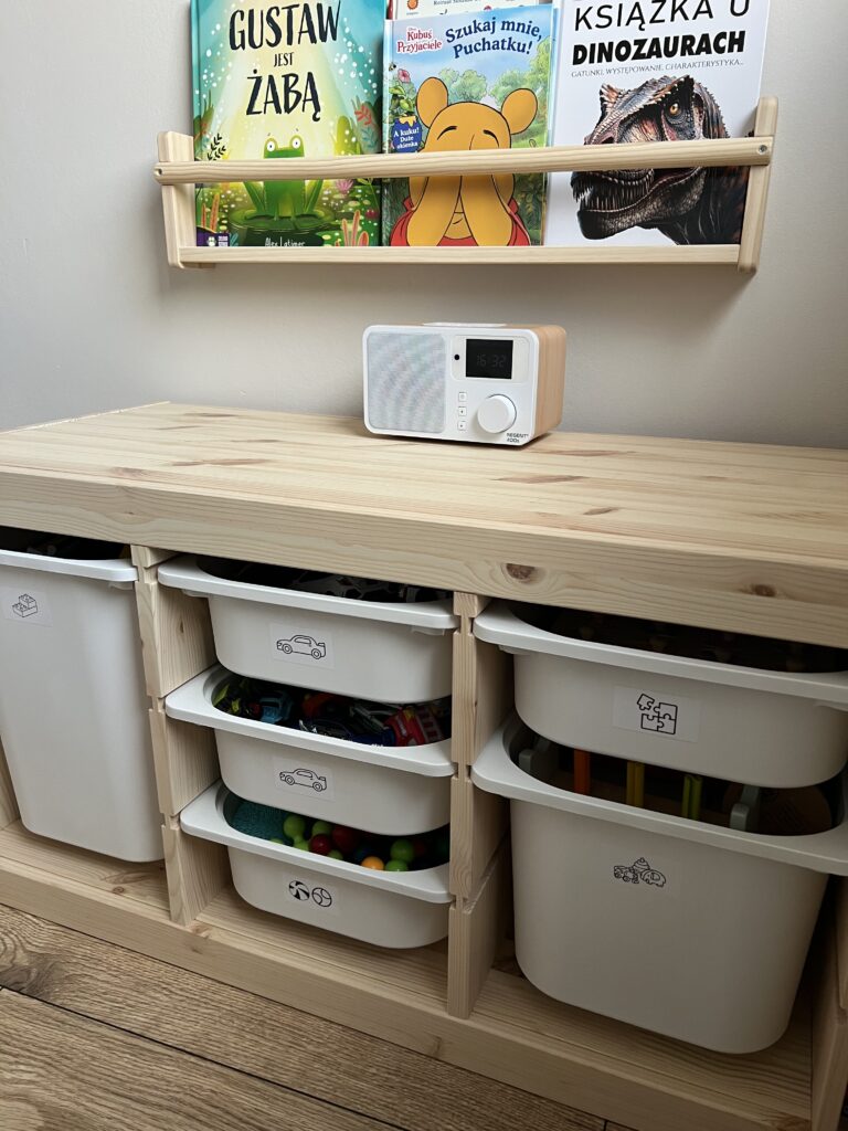 Minimalistyczny, drewniany regał z plastikowymi, wysuwanymi koszami na zabawki w pokoju dziecięcym.